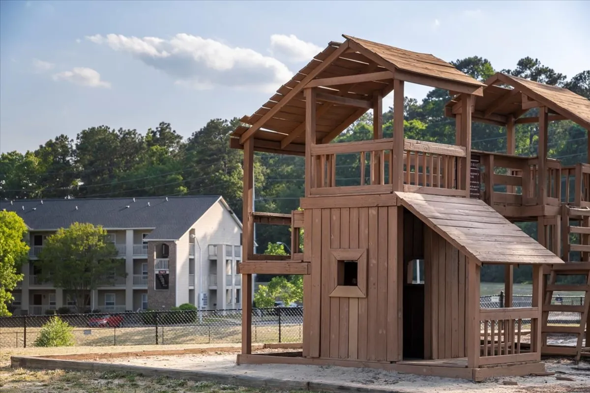 A cheerful outdoor playground set among mature trees and lawns, with a residential building visible in the background. The scene is lively, suggesting children playing, family picnics, and neighbors gathering for community fun.