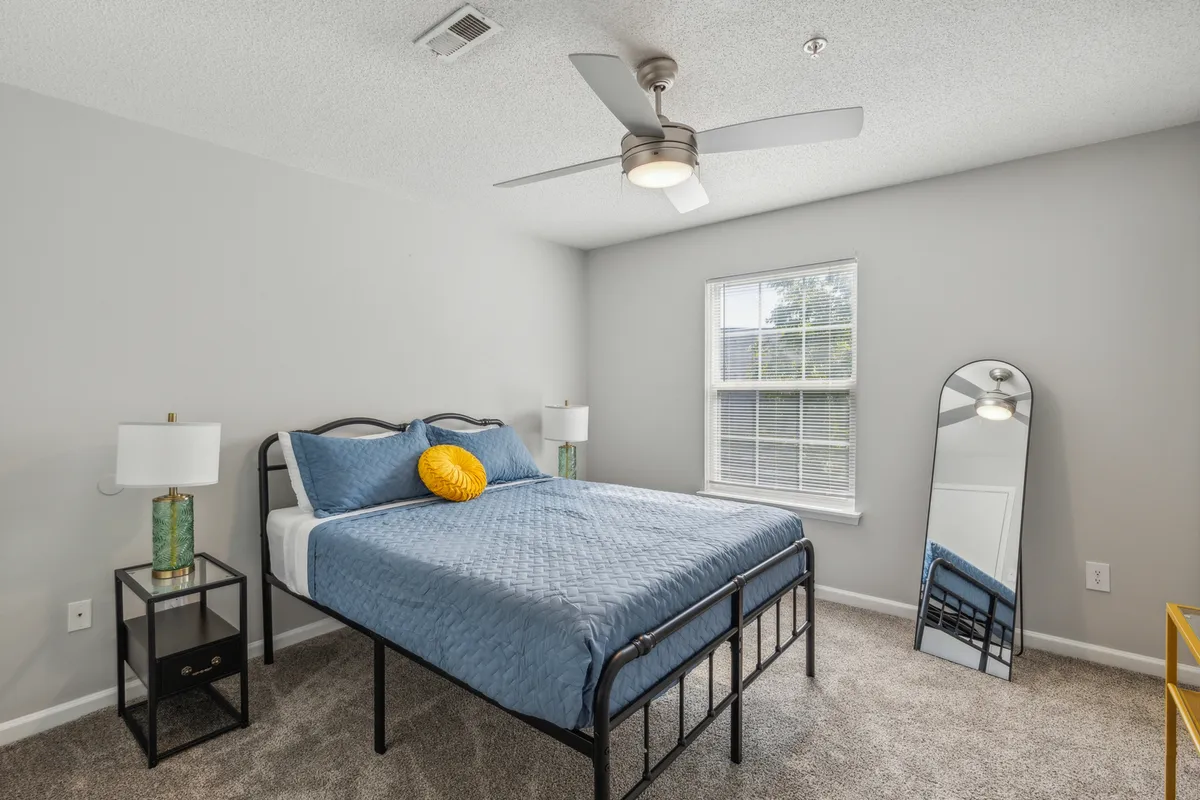 A serene bedroom scene with blue bedding, a yellow accent pillow, full-length mirror, and nightstands with lamps. The room is well-lit by natural light and styled for a peaceful, comfortable ambiance.