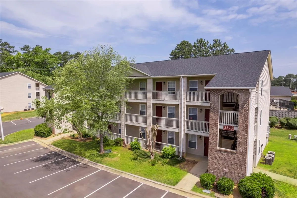 A three-story apartment building surrounded by well-maintained green landscaping and ample parking spaces. The scene conveys a sense of community and ease of access, with walkways and common areas visible for outdoor relaxation and activities.