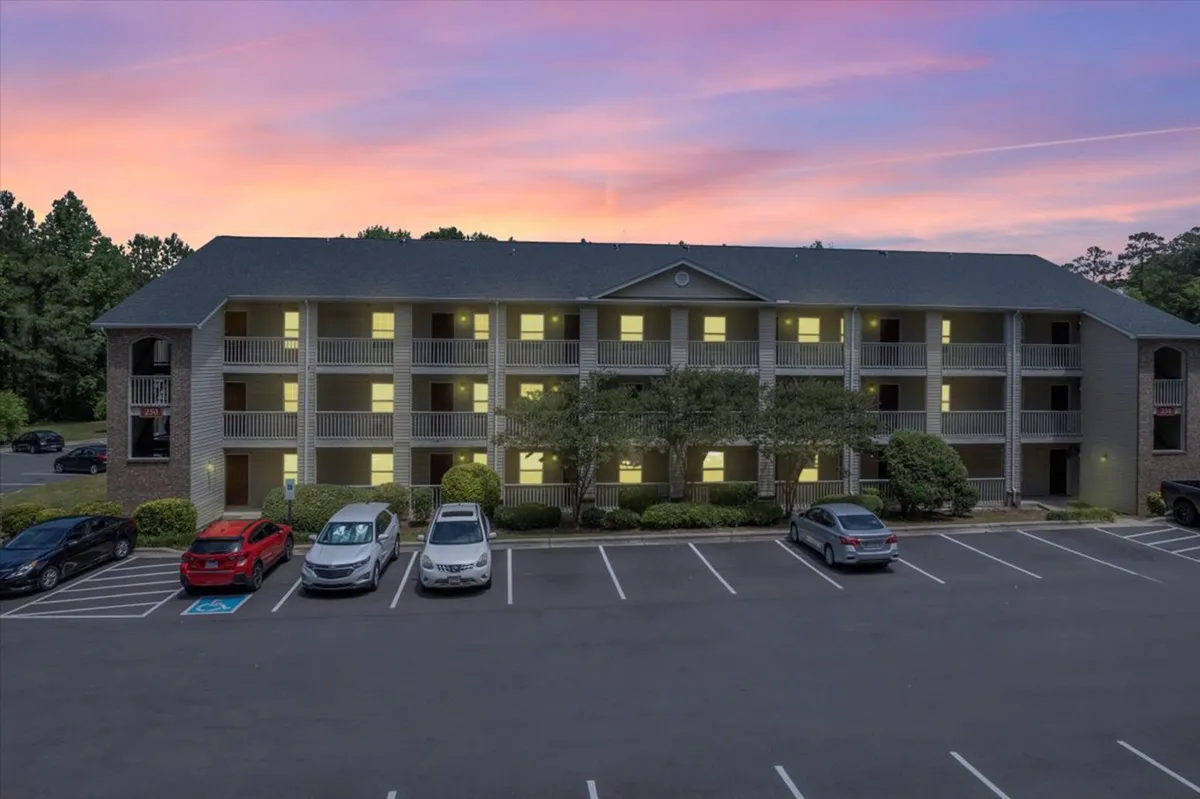 An apartment building with a welcoming facade and ample parking, set against a dramatic sunset sky. Residents are seen socializing on balconies and relaxing outdoors, adding warmth and a sense of community to the scene.
