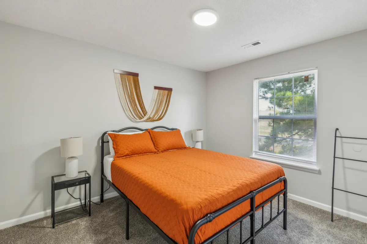 This image features a cozy bedroom with an orange bedspread, a large window letting in natural light, a decorative wall hanging above the bed, and a nightstand with a lamp. The space feels inviting and restful, clearly designed for comfort and relaxation.
