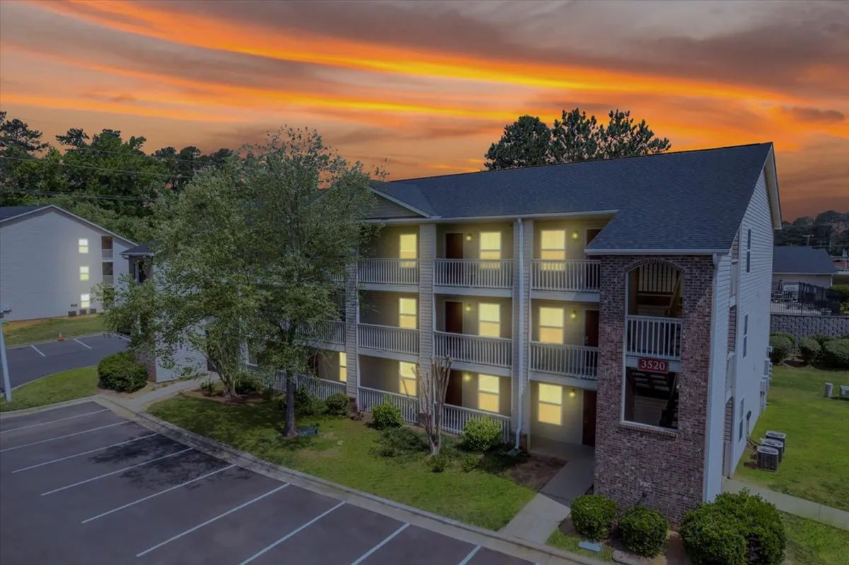 A modern apartment building set against a striking sunset sky, framed by lush greenery. Residents can be seen strolling through the parking area and relaxing on balconies, highlighting both the ease of access and the relaxing atmosphere.