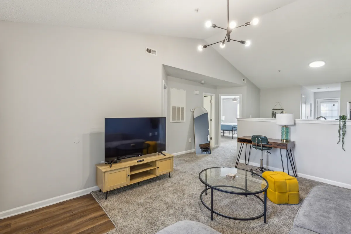 A cozy living room set up with a comfortable sofa, a yellow ottoman, a coffee table, a modern lamp, and a television. The layout is ideal for watching movies, entertaining guests, or simply unwinding, with a contemporary aesthetic that feels welcoming and homey.