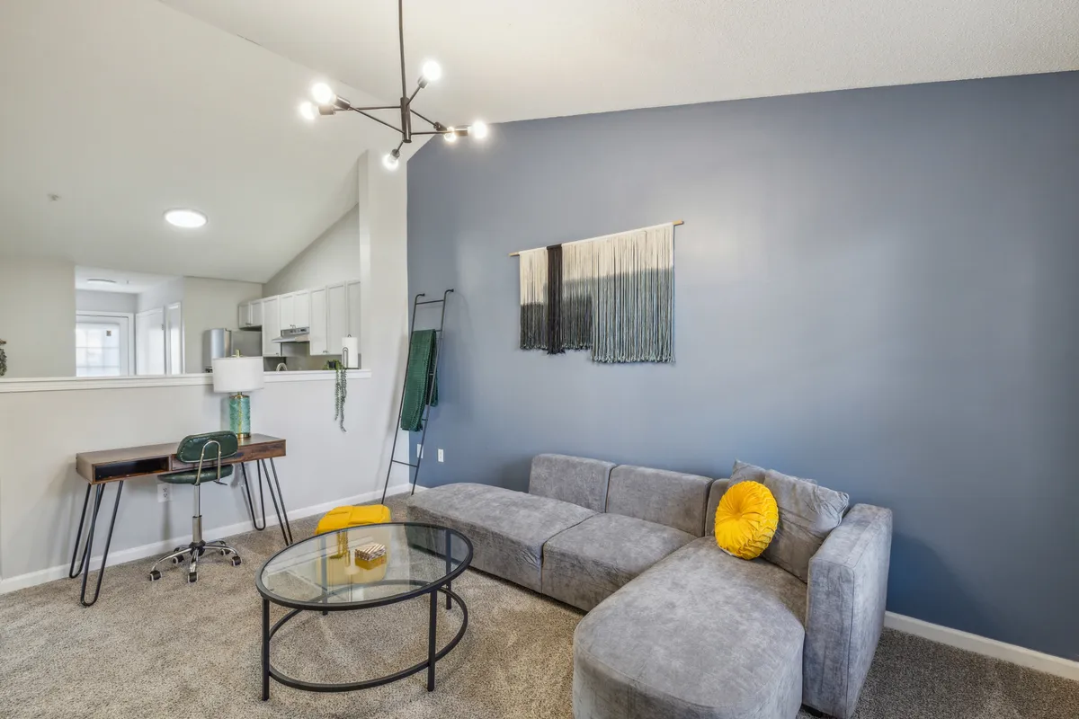 A spacious open-plan living room featuring a contemporary gray sectional sofa, a glass coffee table, and a colorful wall hanging. The area also includes a desk with a chair by the window, allowing for both relaxation and productive work. Abundant natural light streams in, enhancing the feeling of comfort and warmth.