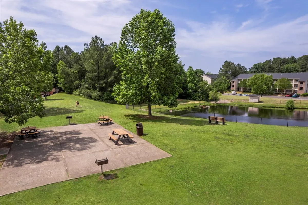 A serene setting with picnic tables under tall green trees beside a peaceful pond, benches for relaxing, and space for barbecues or reading outdoors. The scene is calm, with people enjoying picnics and quiet time in nature.