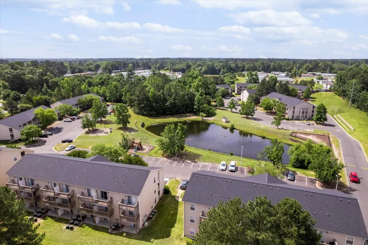 A scenic exterior shot highlighting a tranquil pond ringed by lush green spaces, with modern apartment buildings in the background and a well-equipped playground visible to the side. People are seen walking and children are playing, capturing a lively community atmosphere.