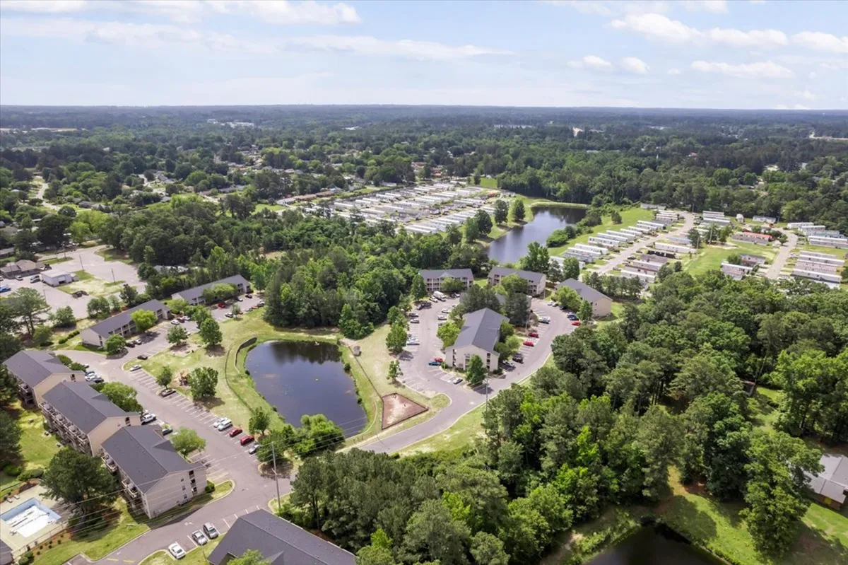A wide outdoor scene featuring lakes surrounded by lush green spaces, with residential buildings and winding roads in the distance. The area is inviting for walking, fishing, picnicking, and biking, and the subtle evening light hints at a peaceful atmosphere perfect for relaxation or community events.