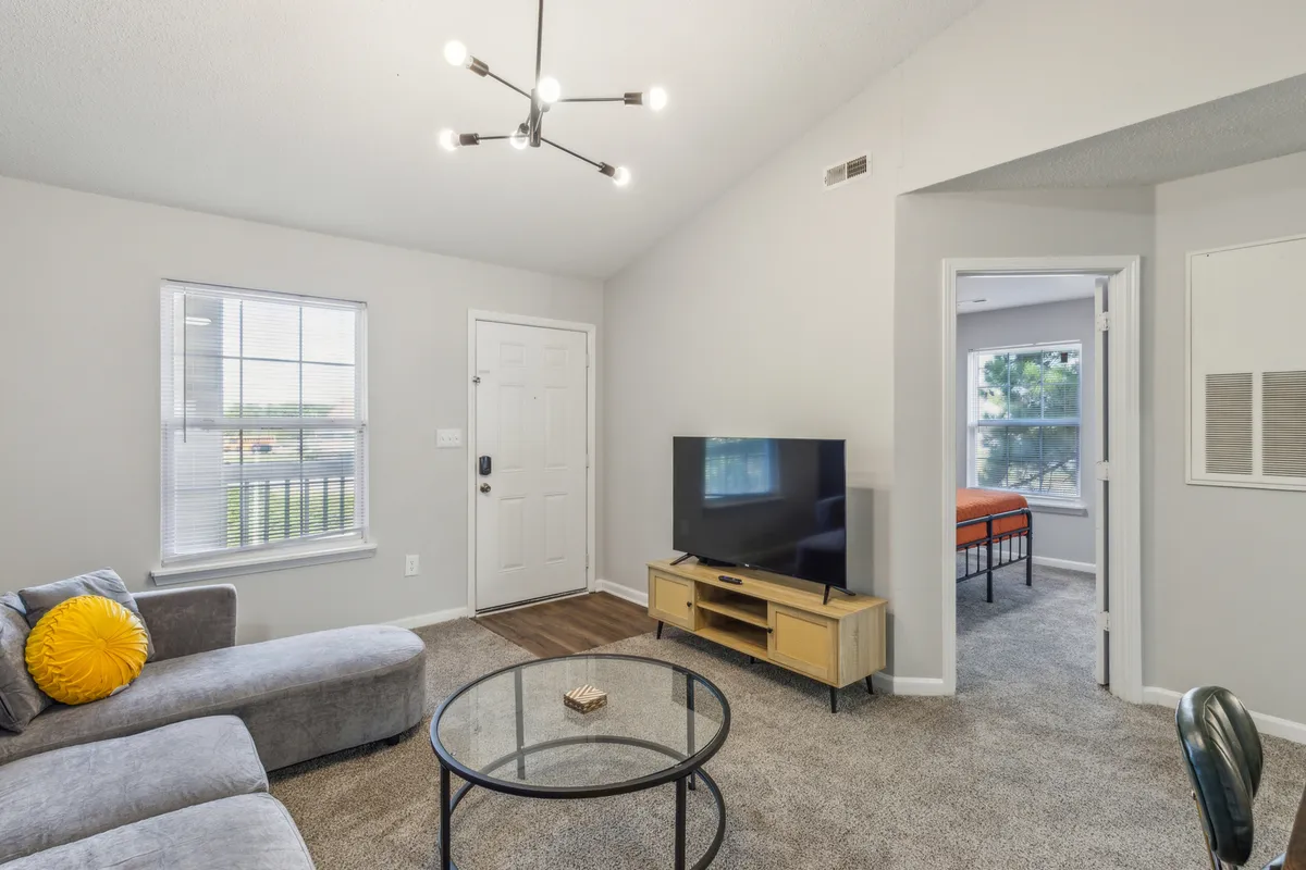 A welcoming living area where a sofa with a yellow accent pillow faces a coffee table and television. The image also captures the open flow between living spaces, with the front door and a doorway to a bedroom visible, suggesting easy movement and a sense of home.